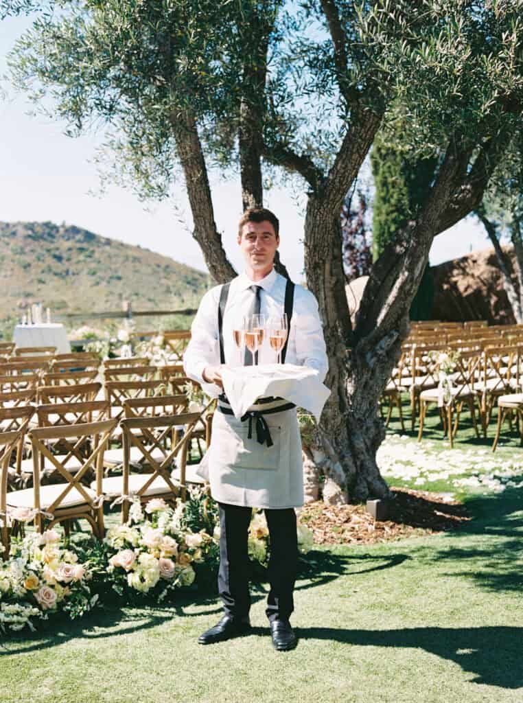 Tres LA Catering waiter holding a tray of champagne glasses, ready to be served to wedding guests before the ceremony. Elegant wedding detail at Cielo Farms in Malibu.