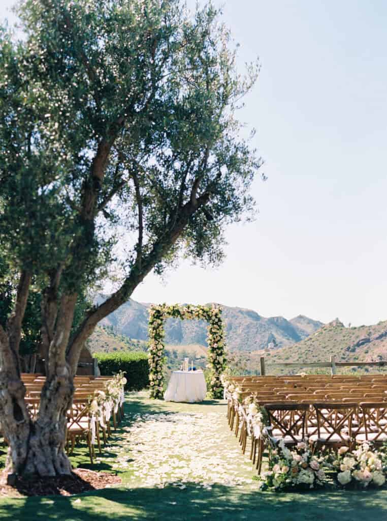 Wedding ceremony setup at Cielo Farms in Malibu, featuring wooden cross-back chairs, a stunning floral arch, and lush floral arrangements with breathtaking views of the Santa Monica Mountains.