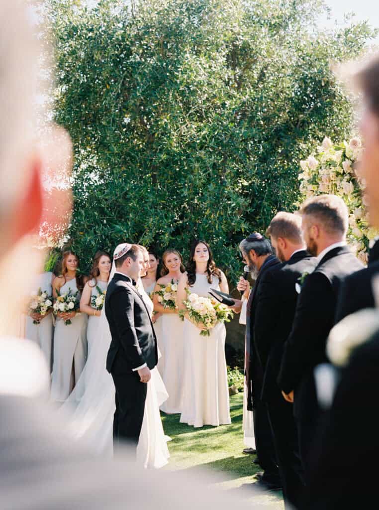 A unique perspective of the bride and groom standing at the altar during their wedding ceremony at Cielo Farms in Malibu, framed by wedding guests looking on with love and admiration.