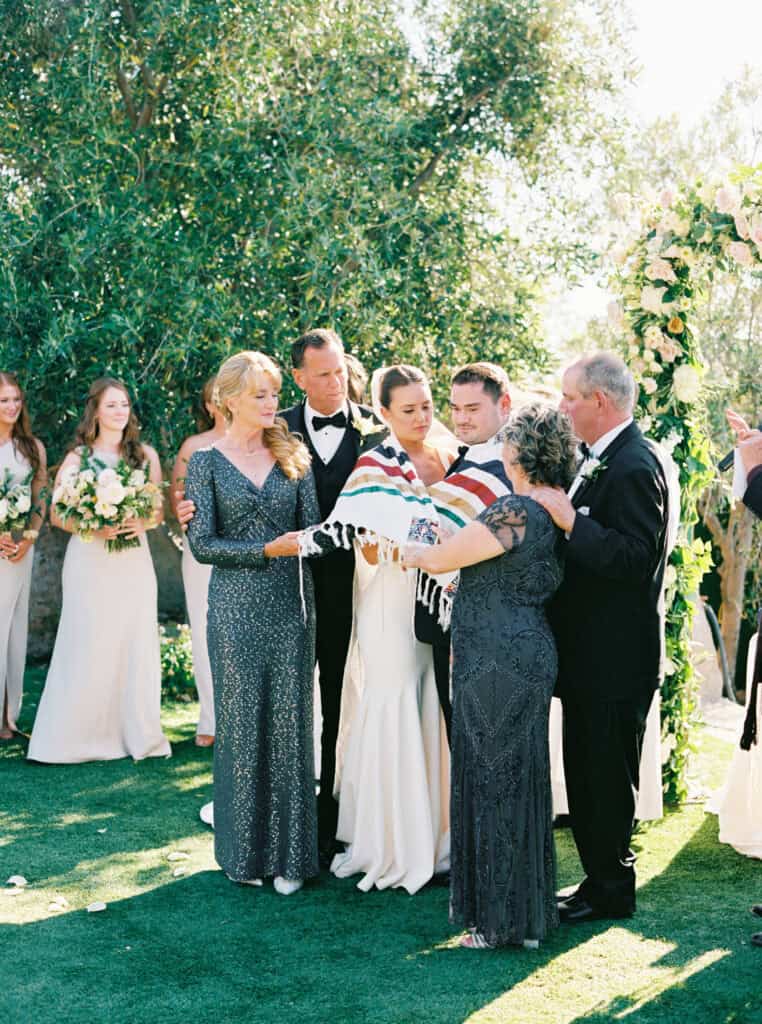 Bride and groom wrapped in a symbolic blanket by their parents during their wedding ceremony at Cielo Farms in Malibu, a heartfelt tradition witnessed by family and friends.