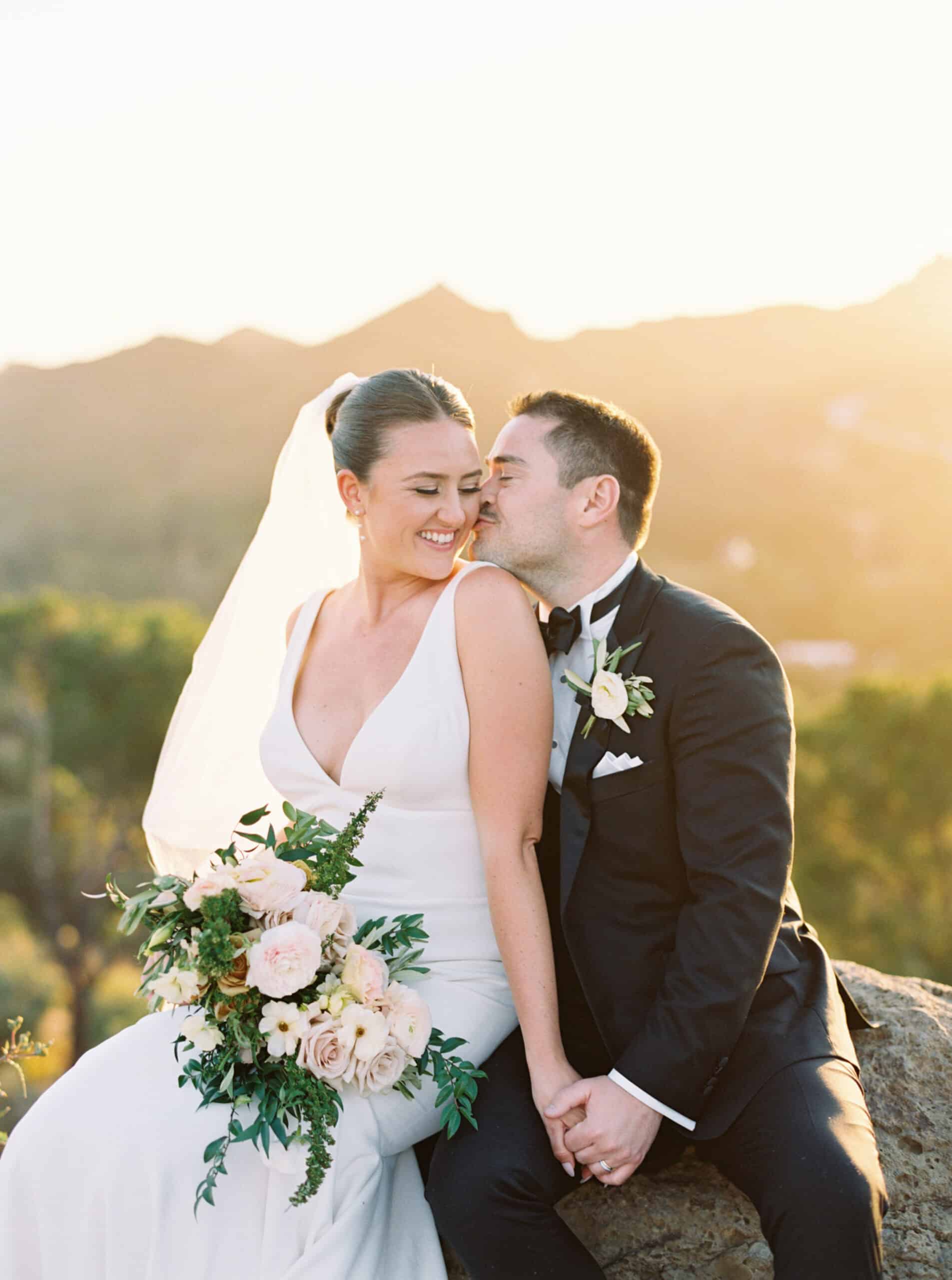 The bride and groom share a sweet moment during their sunset photos at Cielo Farms in Malibu, sitting on a rock as the groom kisses his bride's cheek while she smiles joyfully.