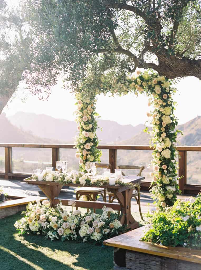 The bride and groom's romantic sweetheart table at Cielo Farms, surrounded by lush florals and a breathtaking floral arch, creating a stunning focal point for their Malibu wedding.