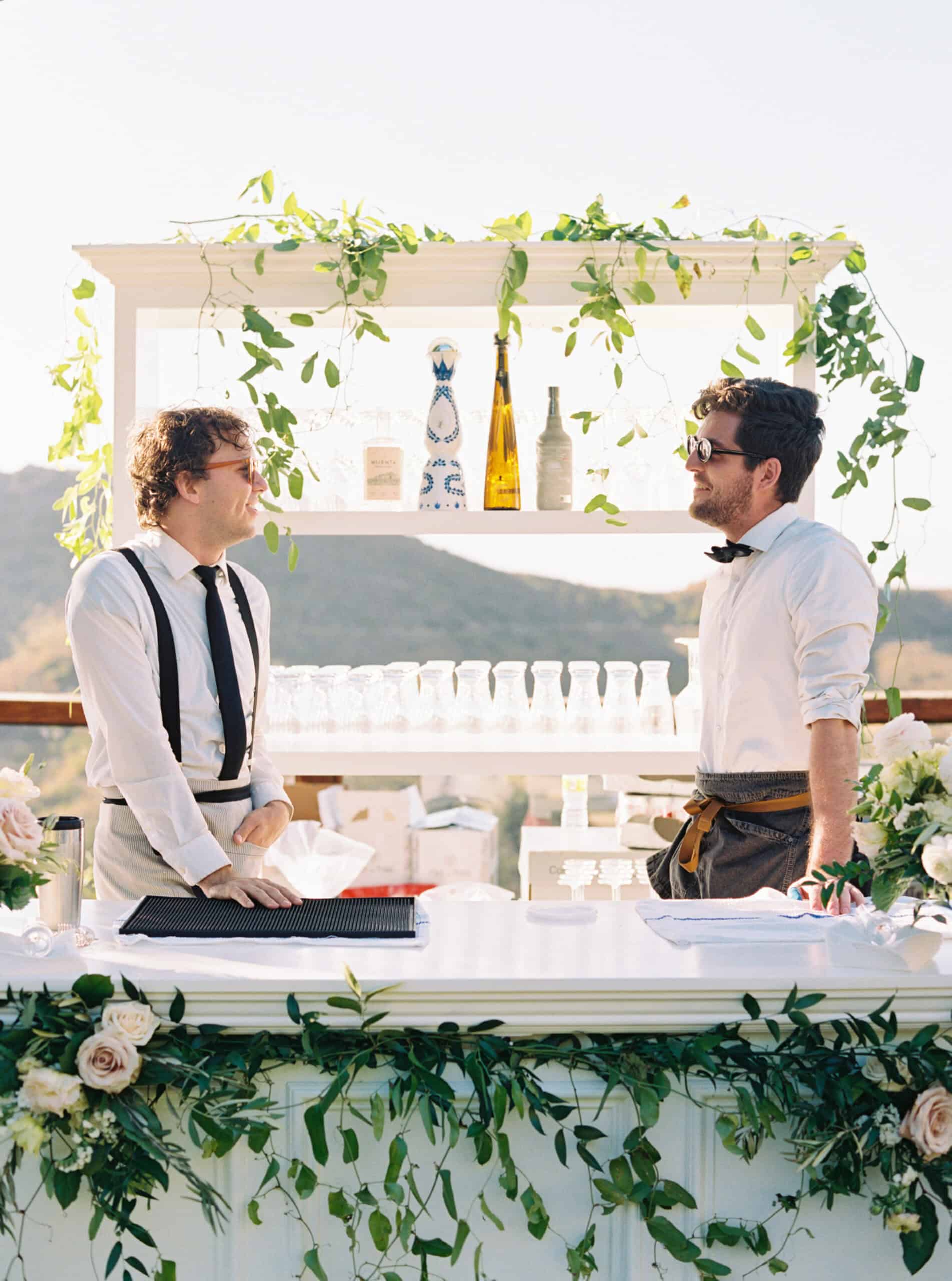 The bride and groom's tequila bar at Cielo Farms, decorated with lush, romantic florals, with two bartenders sharing a moment before guests arrive. Bottles of tequila line the shelves behind them, adding a unique touch to the wedding celebration.
