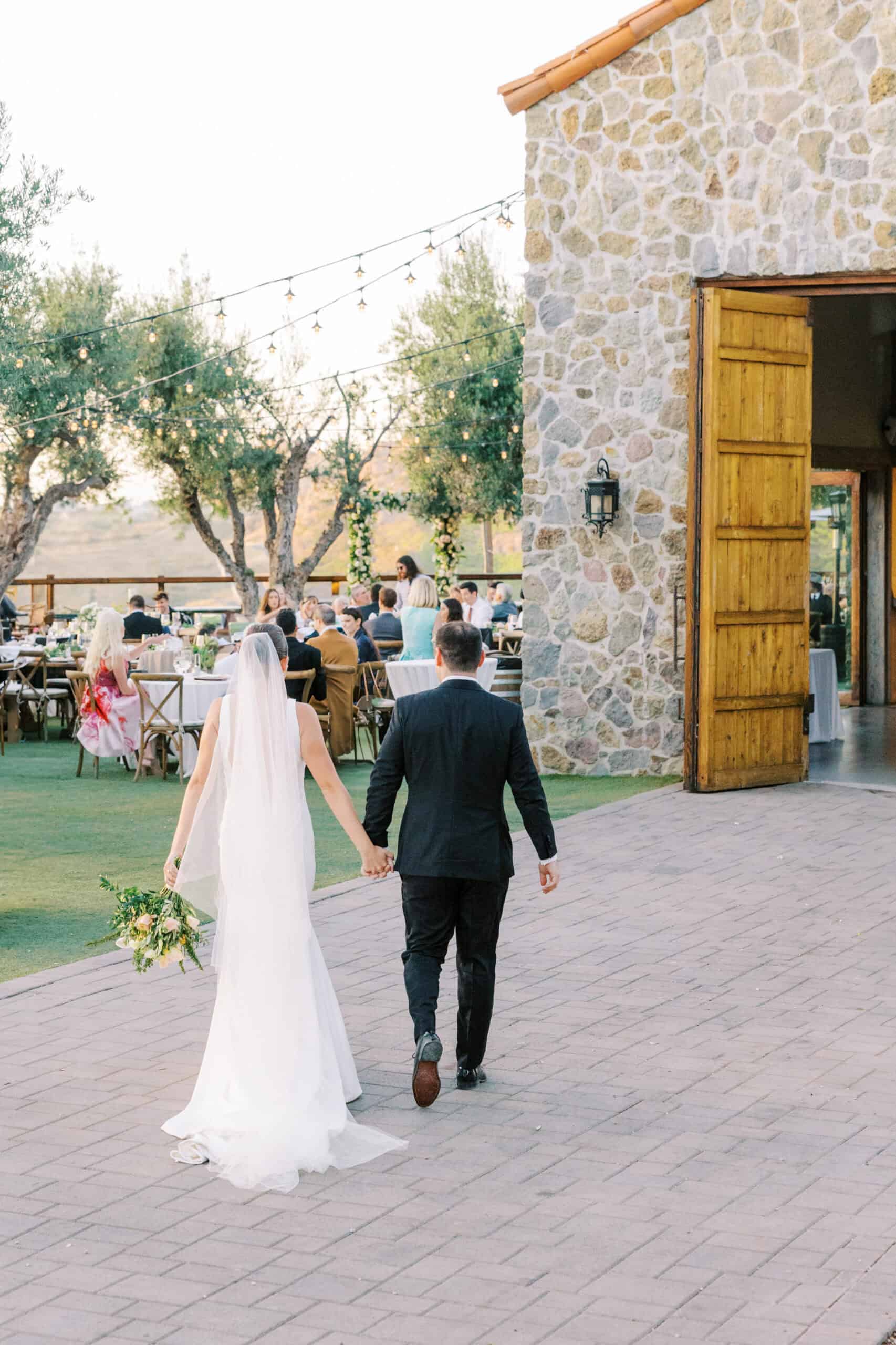 The bride and groom walk back into their reception after sunset photos, with the iconic stone building at Cielo Farms and the elegant reception space in the background, creating a romantic and timeless moment.