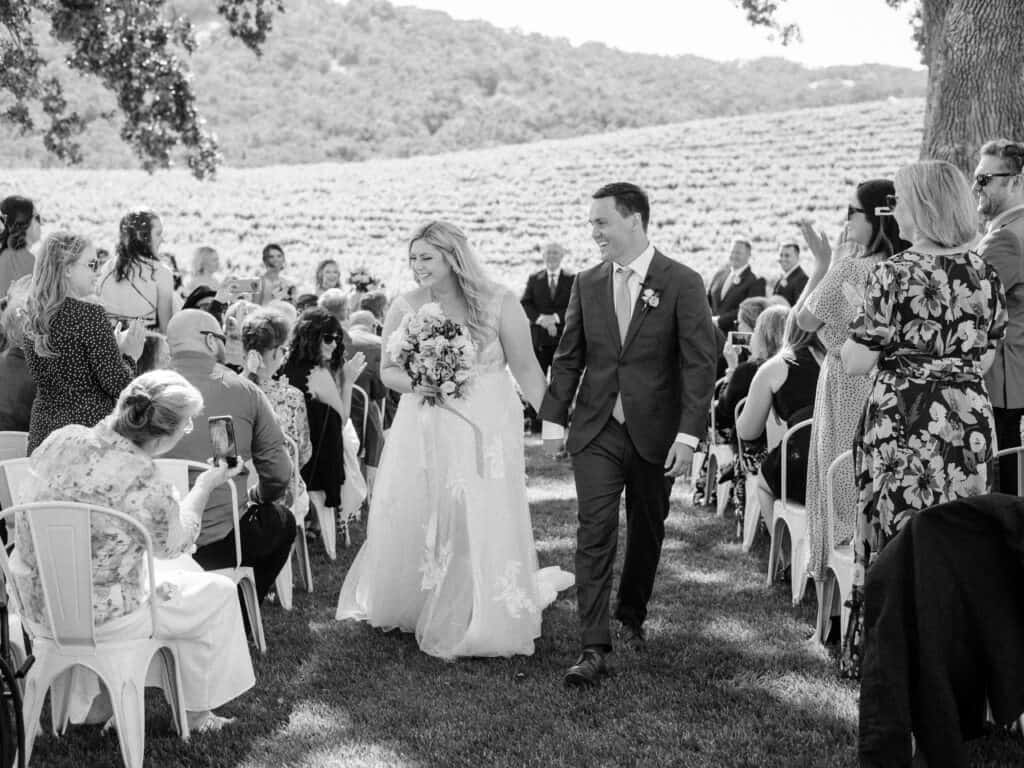 A classic black and white photo of the newlyweds walking back up the aisle at Hammersky Vineyards while their guests cheer in celebration.