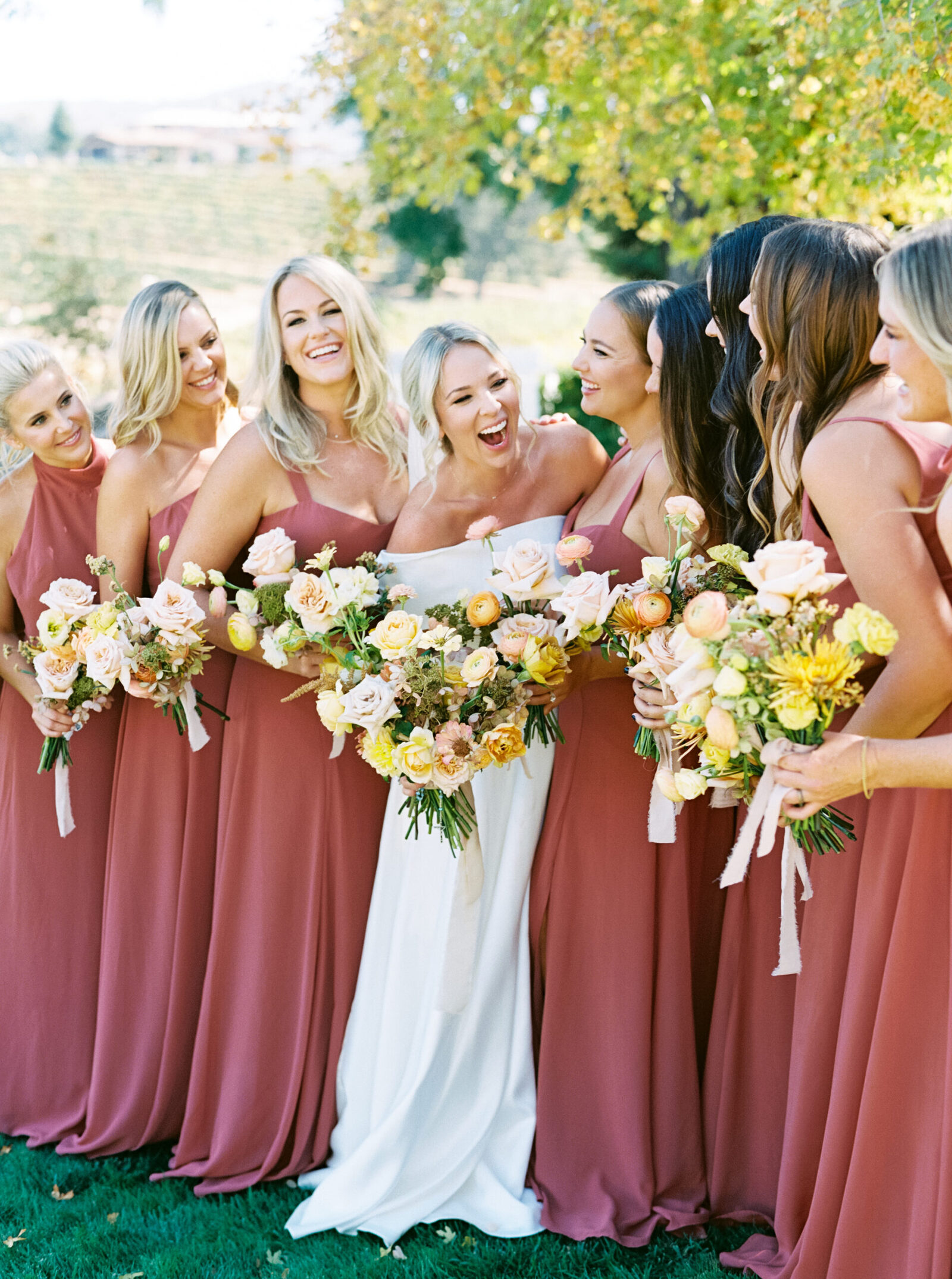 A bride laughs joyfully with her bridesmaids, all holding soft yellow bouquets, at Hammersky Vineyards in Paso Robles.