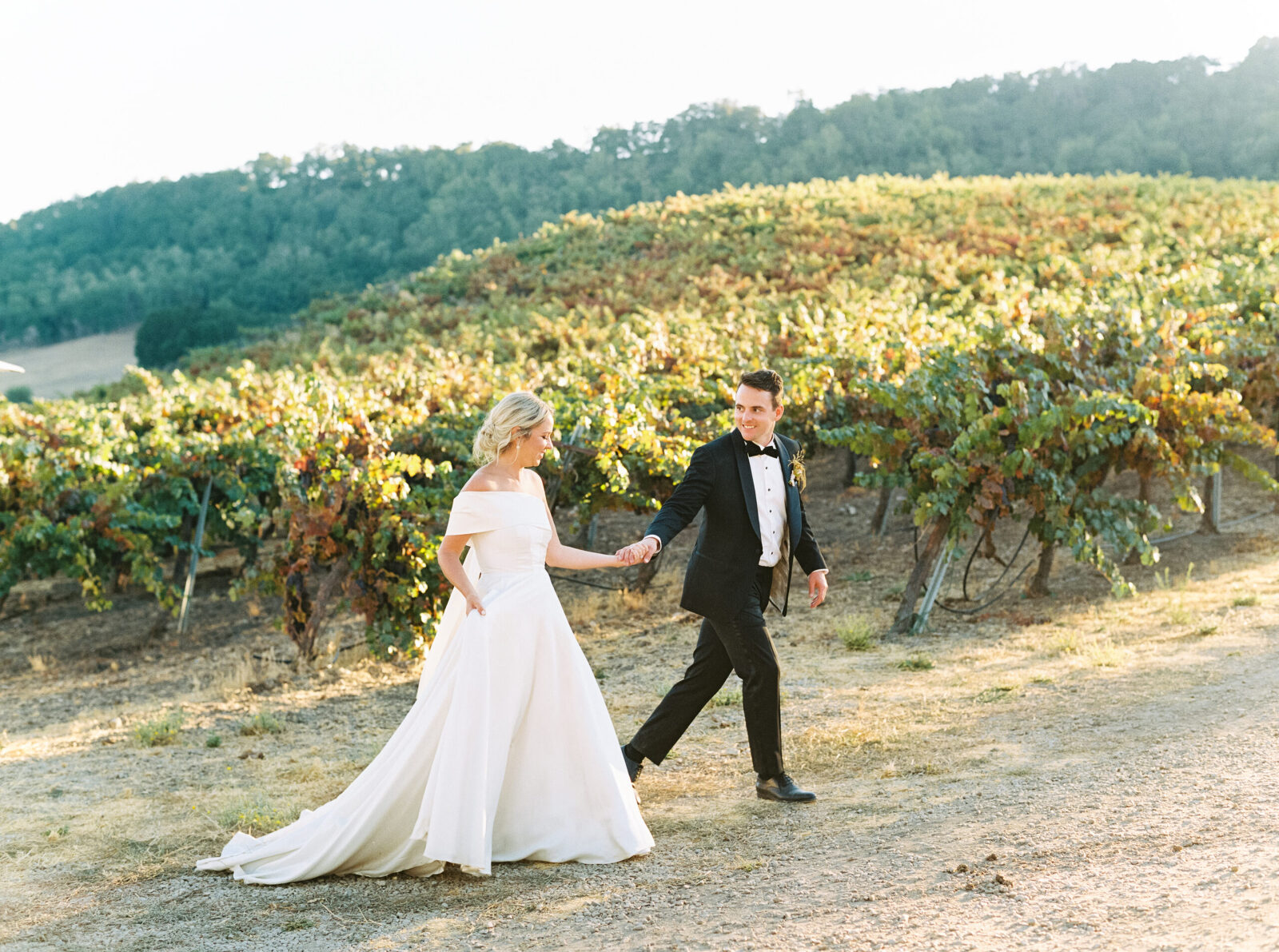 A bride and groom walk hand in hand near the vineyards at Hammersky in Paso Robles, surrounded by rolling hills and golden light.