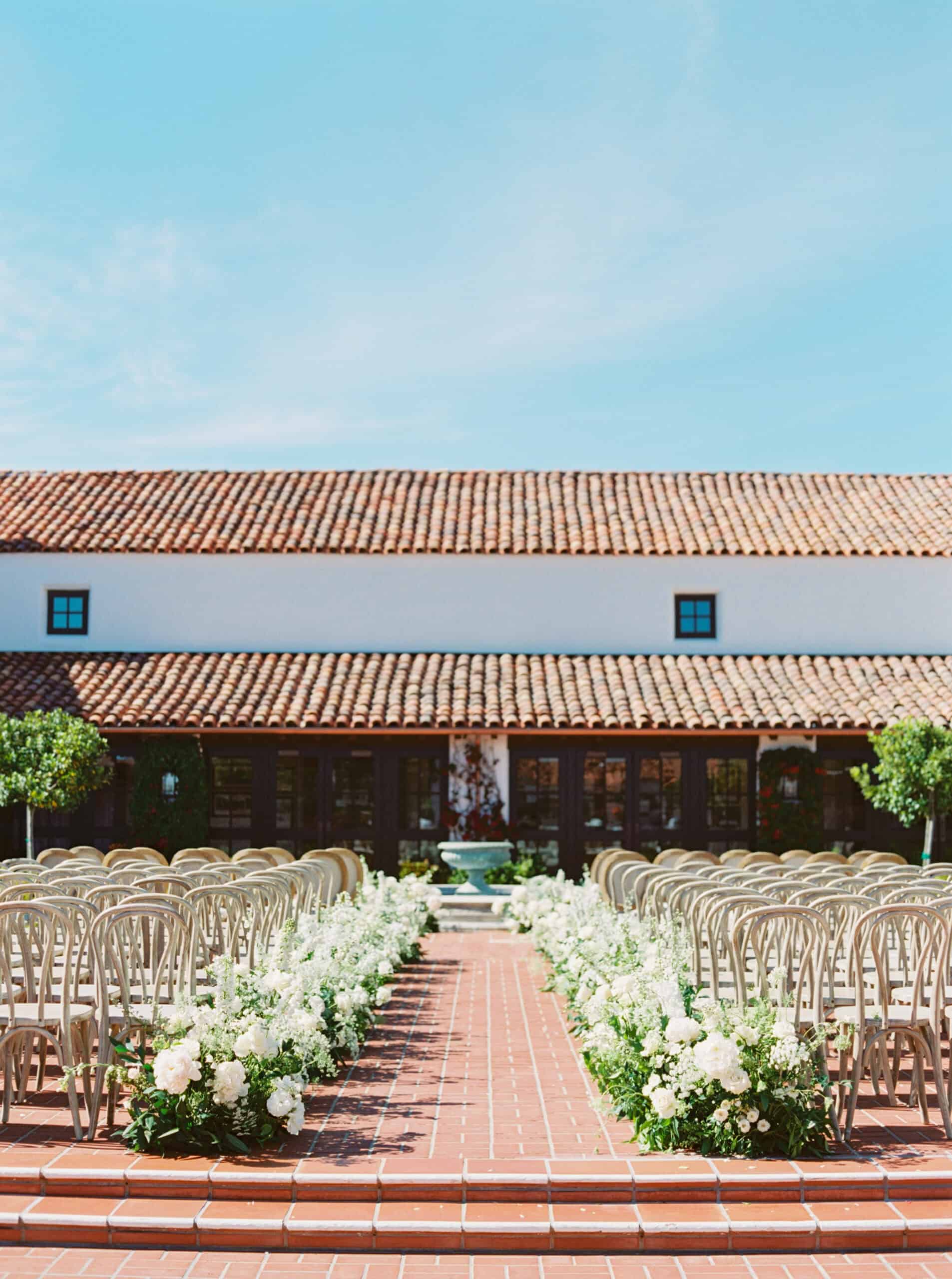 The ceremony location at the Monterey Peninsula Country Club in Pebble Beach, set with whitewashed wooden chairs and white floral arrangements lining the aisle.