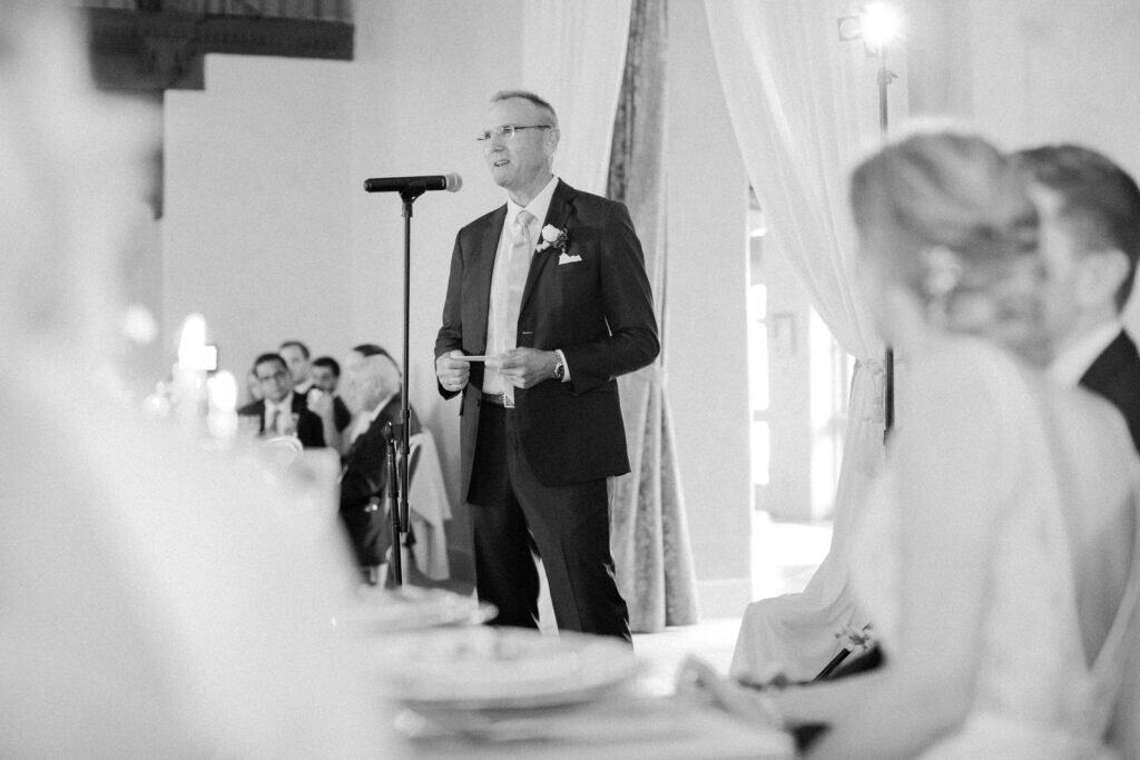 A black and white photo of the bride's father giving a heartfelt speech at the wedding reception, with the bride and groom looking on lovingly.