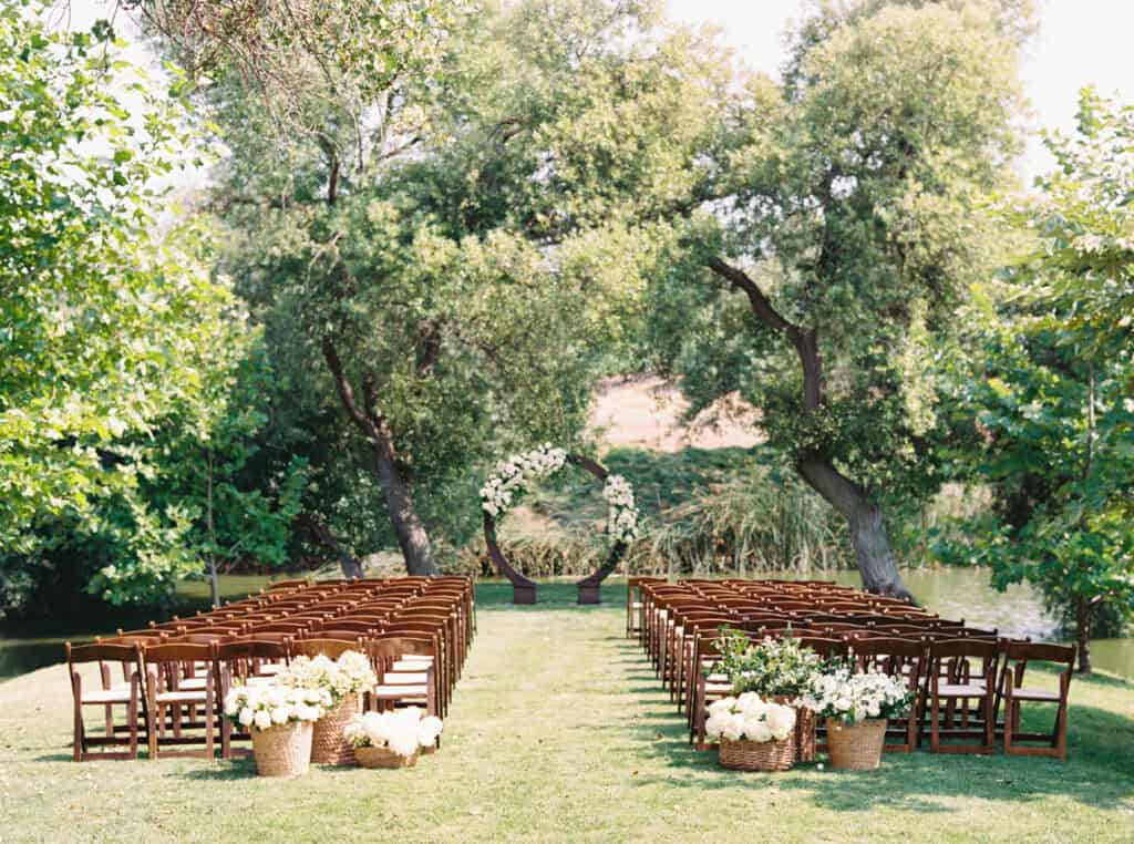 Greengate Ranch wedding ceremony site with wooden chairs arranged by the pond, featuring an elegant floral arch adorned with fluffy white florals and matching baskets at the aisle entrance, set for Michelle and Brendan's romantic celebration.
