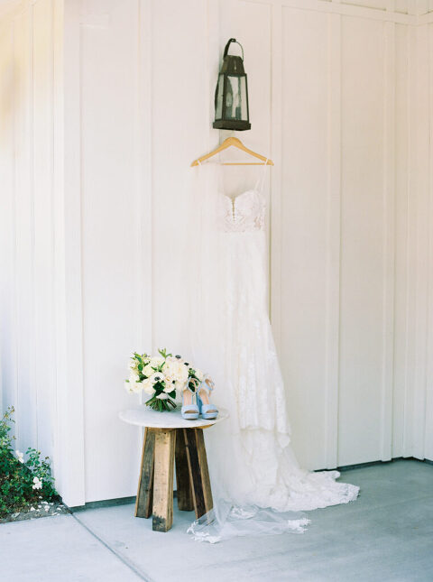 A timeless bridal moment featuring Michelle’s wedding dress hanging gracefully, paired with her classic all-white bouquet and stylish blue suede platform heels. A chic and elegant detail from her Greengate Ranch wedding in San Luis Obispo.