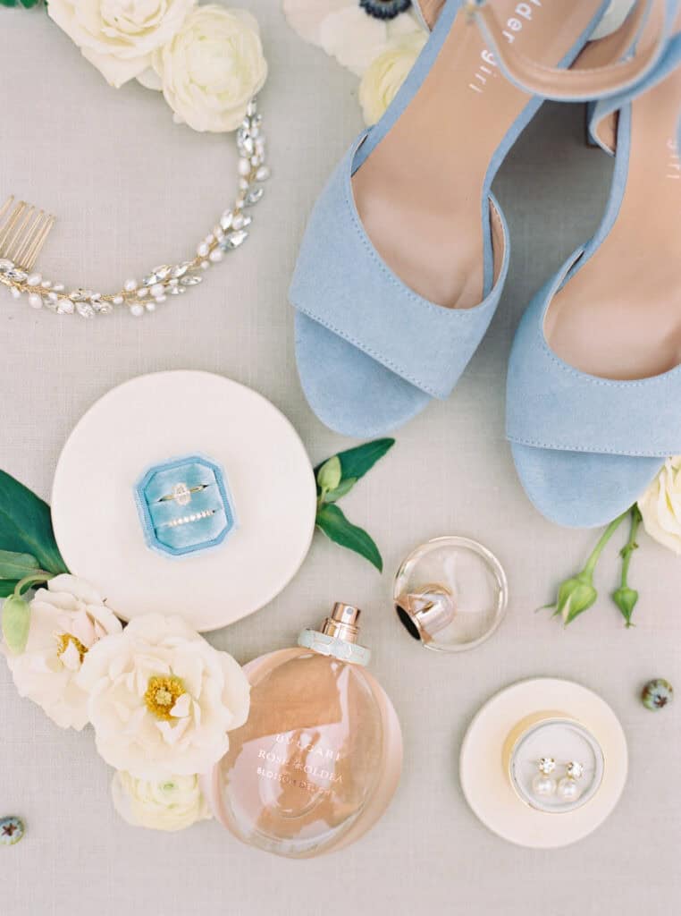 A beautifully styled bridal details flat lay featuring Michelle’s wedding rings, elegant perfume bottle, sparkling earrings, delicate headpiece, and stylish blue suede shoes, accented with fresh white florals for a romantic touch.