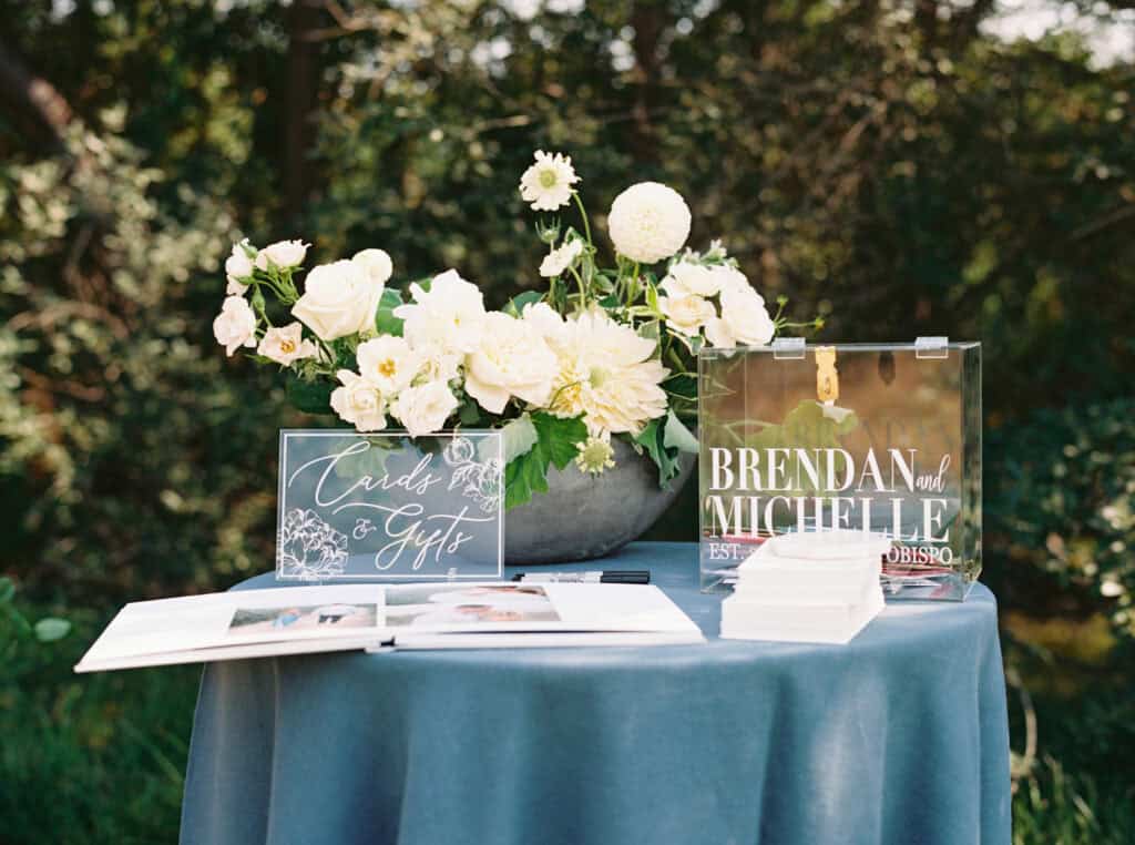 Elegant wedding welcome station with a blue linen-covered table featuring a stunning white floral arrangement, acrylic card box, gift sign, and a guestbook for signing, setting the tone for Michelle and Brendan's celebration.