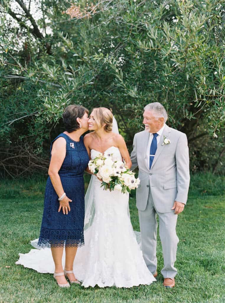 Emotional family photo of Michelle sharing a tender moment with her parents as her mom gives her a sweet kiss on the cheek, capturing the love and connection during their Greengate Ranch wedding.