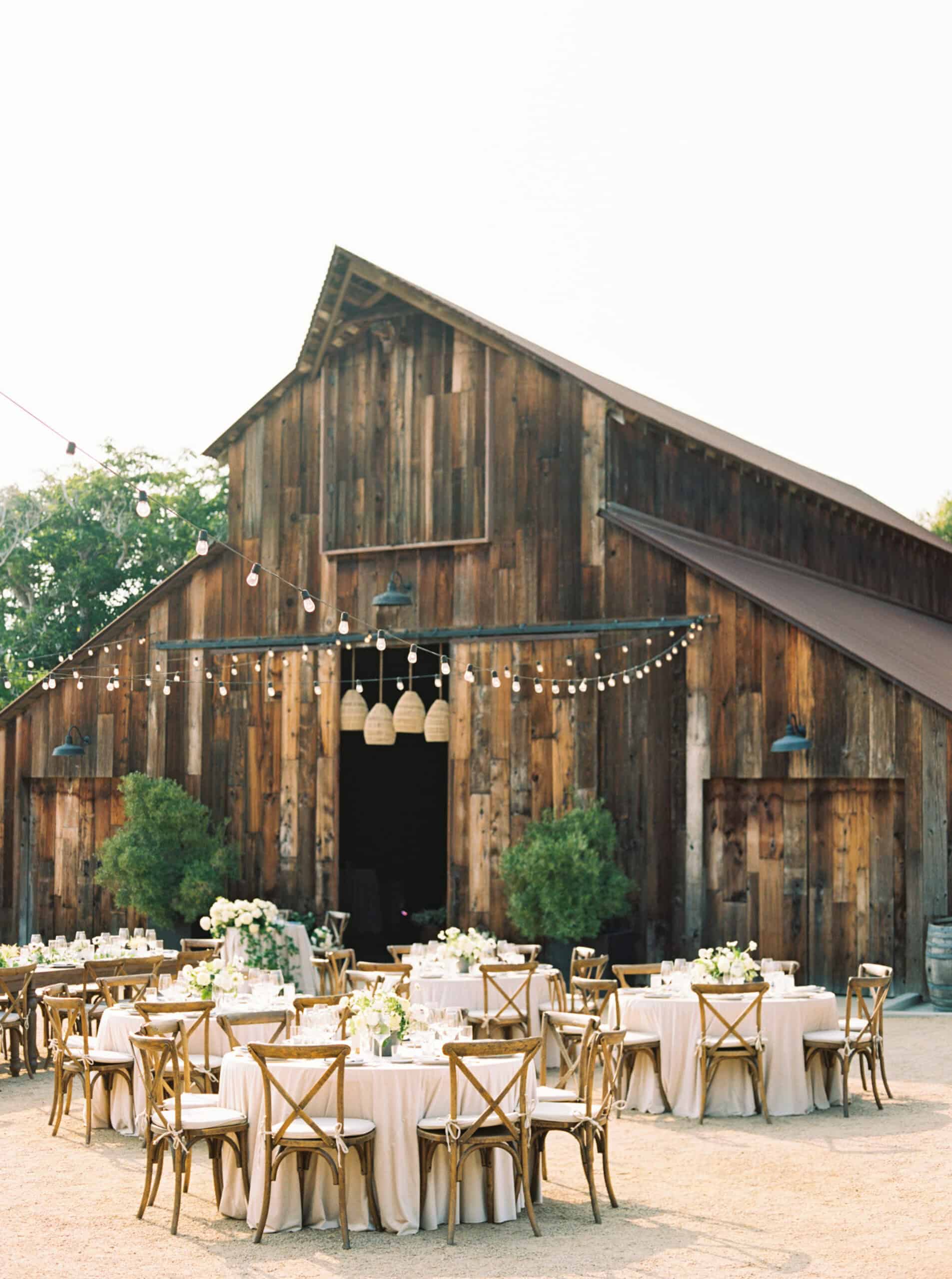 Iconic shot of the stunning barn at Greengate Ranch, beautifully set for guests with soft pink linens, wooden cross-back chairs, and elegant all-white floral centerpieces, creating a dreamy wedding reception setting.