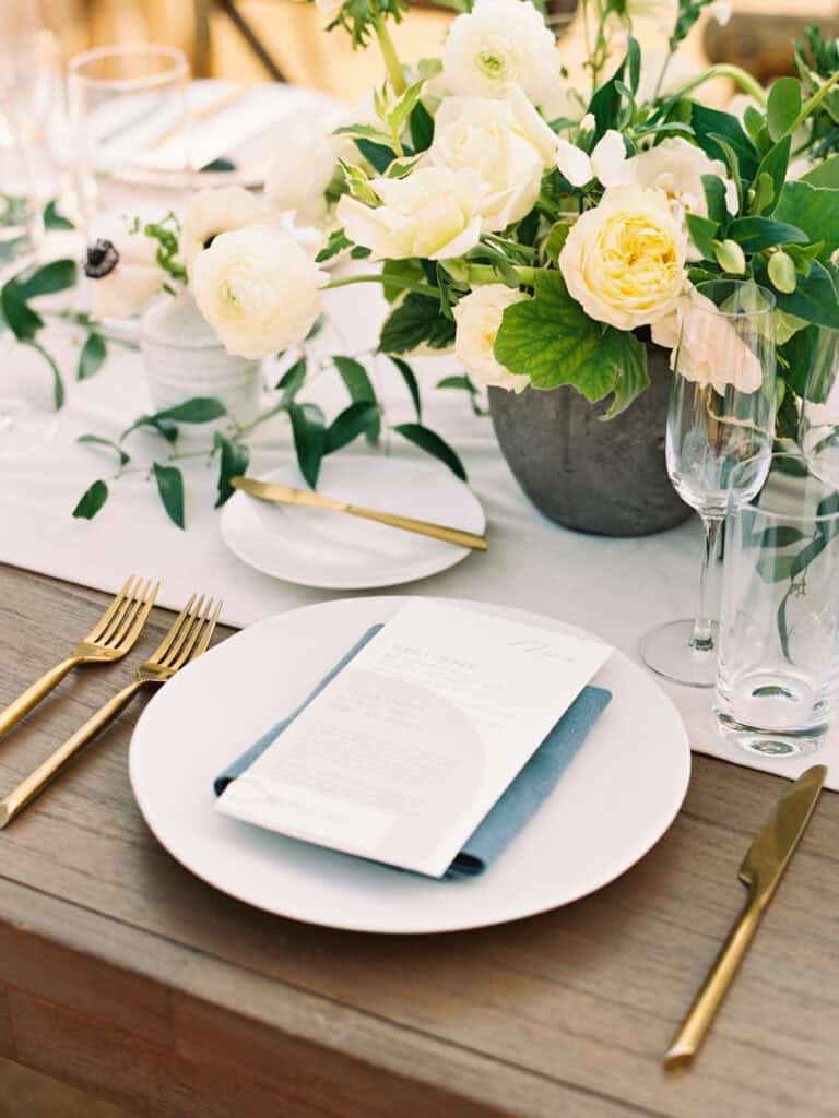 Timeless wedding place setting featuring white plates, blue cloth napkins, custom menus by Swell Press, and elegant white floral arrangements by Noonan's Designs, creating a sophisticated dining experience at Greengate Ranch.