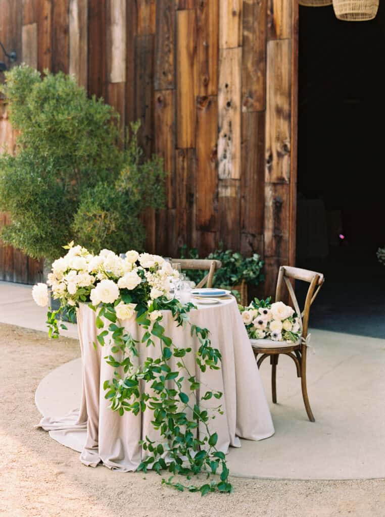 Romantic sweetheart table set for the newlyweds at Greengate Ranch, featuring soft pink linens, a cascading white floral arrangement, and charming wooden cross-back chairs, creating a perfect intimate setting.