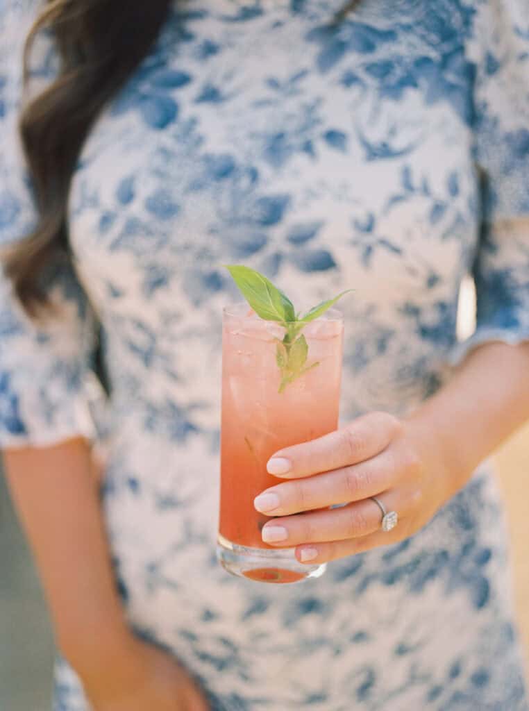 Guest enjoying a pink signature drink at Michelle and Brendan's wedding, with a stunning blue floral dress providing a beautiful backdrop, adding to the vibrant cocktail hour atmosphere at Greengate Ranch.