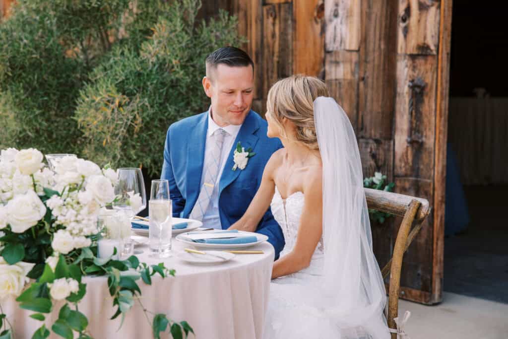 Michelle and Brendan sit at their romantic sweetheart table at Greengate Ranch, with Brendan adoringly looking at his wife, capturing a tender moment during their wedding celebration.