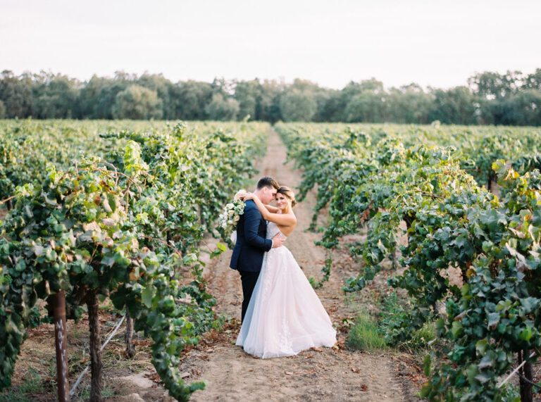 A romantic film photo of newlyweds posing in the vineyards at sunset at a private estate in Lodi, CA. The bride wraps her arms around her groom’s neck, smiling at the camera while he snuggles into her.