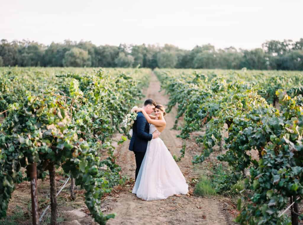 A romantic film photo of newlyweds posing in the vineyards at sunset at a private estate in Lodi, CA. The bride wraps her arms around her groom’s neck, smiling at the camera while he snuggles into her.