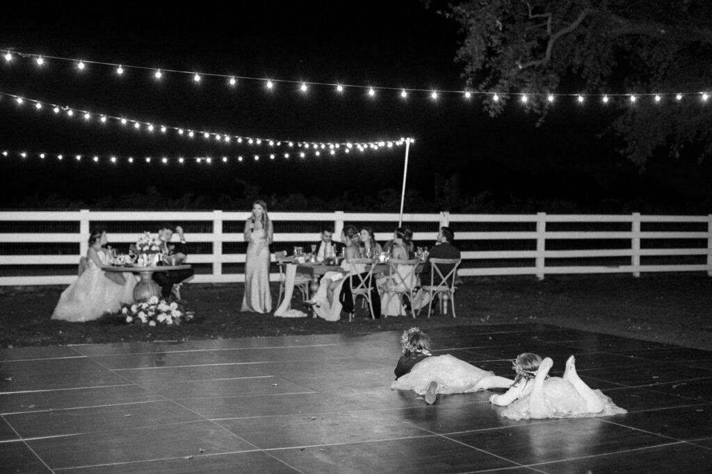 Two flower girls laying on the ground, watching the maid of honor give her heartfelt wedding speech during the ceremony.