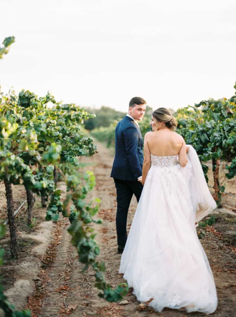 The groom leads his bride through the vineyards, looking back at her with love at their private estate wedding in Lodi, CA.