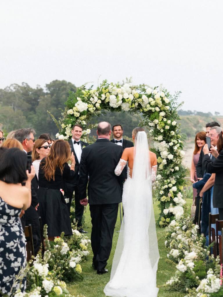 A bride walking down the aisle with her father towards her future husband on the ceremony lawn at the Bacara Resort. 