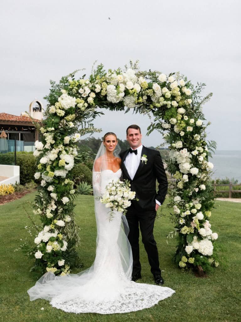 Stunning portrait of a bride and groom under a lush white and green floral arch at their wedding ceremony at the Bacara Resort in Santa Barbara, capturing a romantic and elegant moment.
