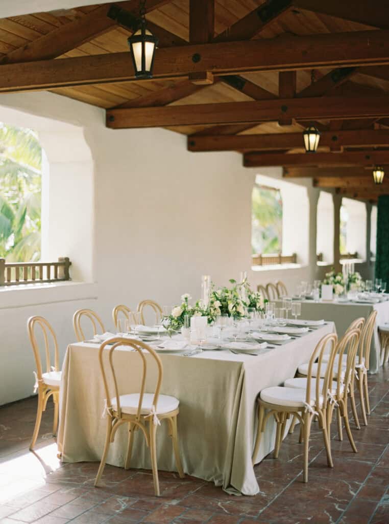 Full wedding reception table set with soft neutral tones, classic white floral arrangements, light wooden chairs, and flickering candles, creating an elegant and timeless atmosphere for guests to enjoy at the Ritz Carlton Bacara Resort. 