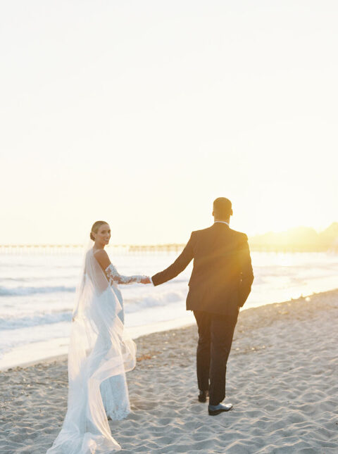 Newlyweds walking hand in hand along the beach at sunset, with the stunning Bacara Resort in Santa Barbara as the backdrop, capturing the perfect romantic moment on their luxury wedding day.