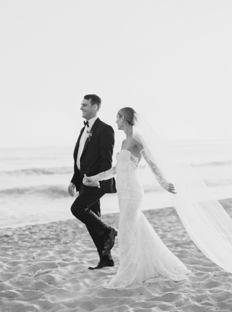A fun black and white photo of the newlyweds walking hand in hand along the beach, with the bride's veil flowing gracefully in the breeze, capturing a moment of joy and love.