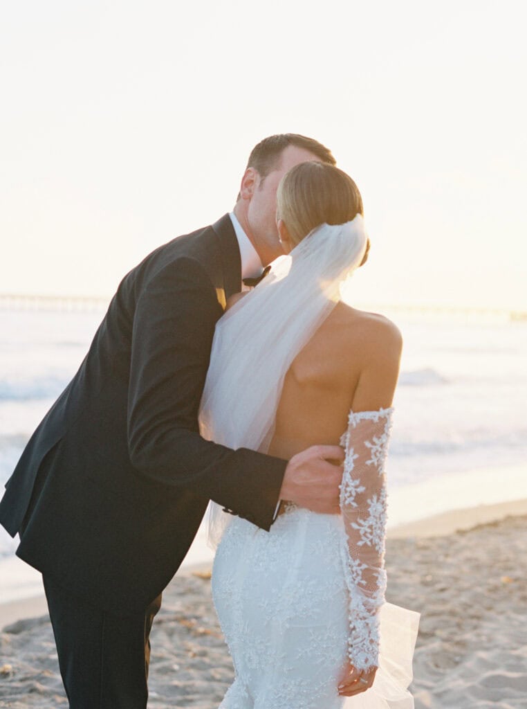 A groom kisses his bride during a romantic sunset photo session on the beach at the Ritz Carlton Bacara Resort, capturing a moment of love and tranquility.