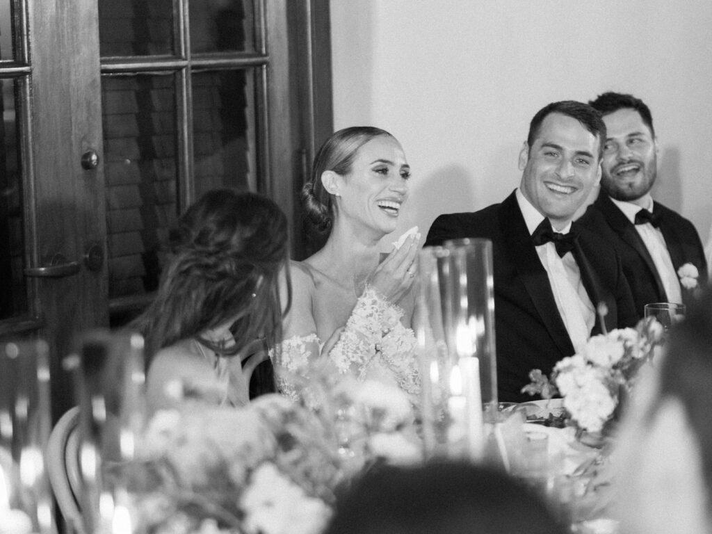 The bride listens with joy and emotion during her wedding toasts, smiling through tears as she takes in the heartfelt words from her loved ones.